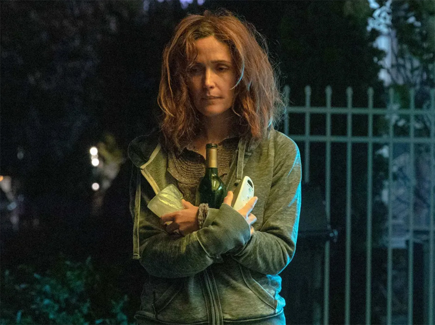 A frowning woman in a hoodie crosses her arms, holding a wine bottle and two other items, standing outside in a backyard